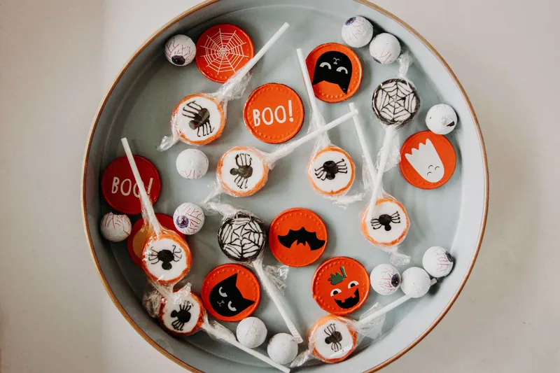 A shallow bowl of Halloween lollipops with orange and white spider, bat, and "BOO" designs, seen from above on a soft neutral background.